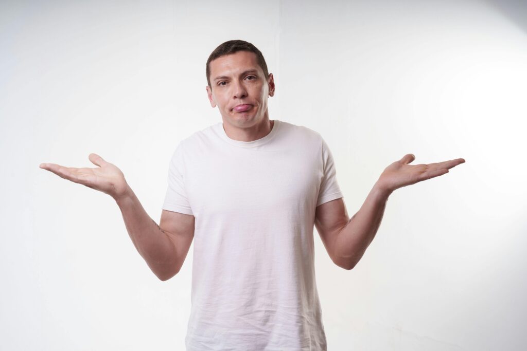 Adult man in white t-shirt shrugs in studio, expressing confusion against a plain background.